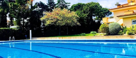 A clear blue swimming pool with a diving board, surrounded by trees and a building with a tiled roof.