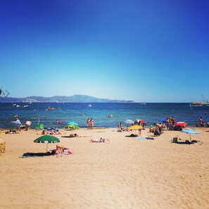 Uma praia com pessoas tomando sol e nadando, guarda-sóis e barcos na água.