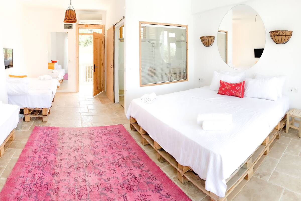 A hotel room with a large bed, a smaller bed, a pink patterned rug, wooden pallets, a large mirror, and a wooden door.