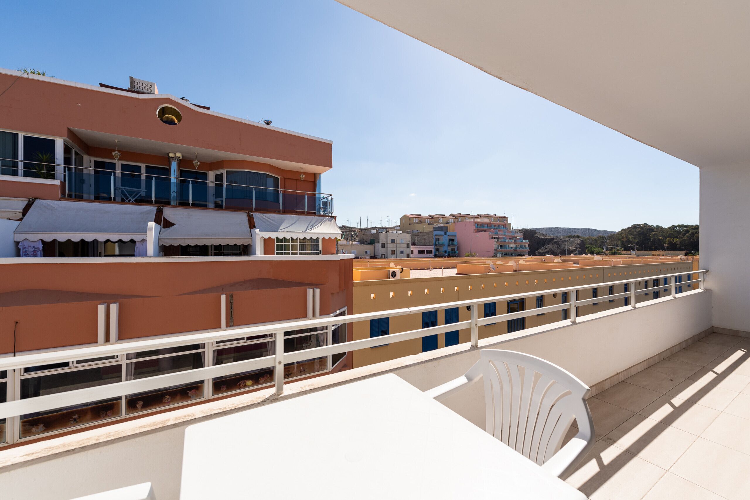 Un balcón con una silla blanca, barandilla y vista de edificios al fondo.