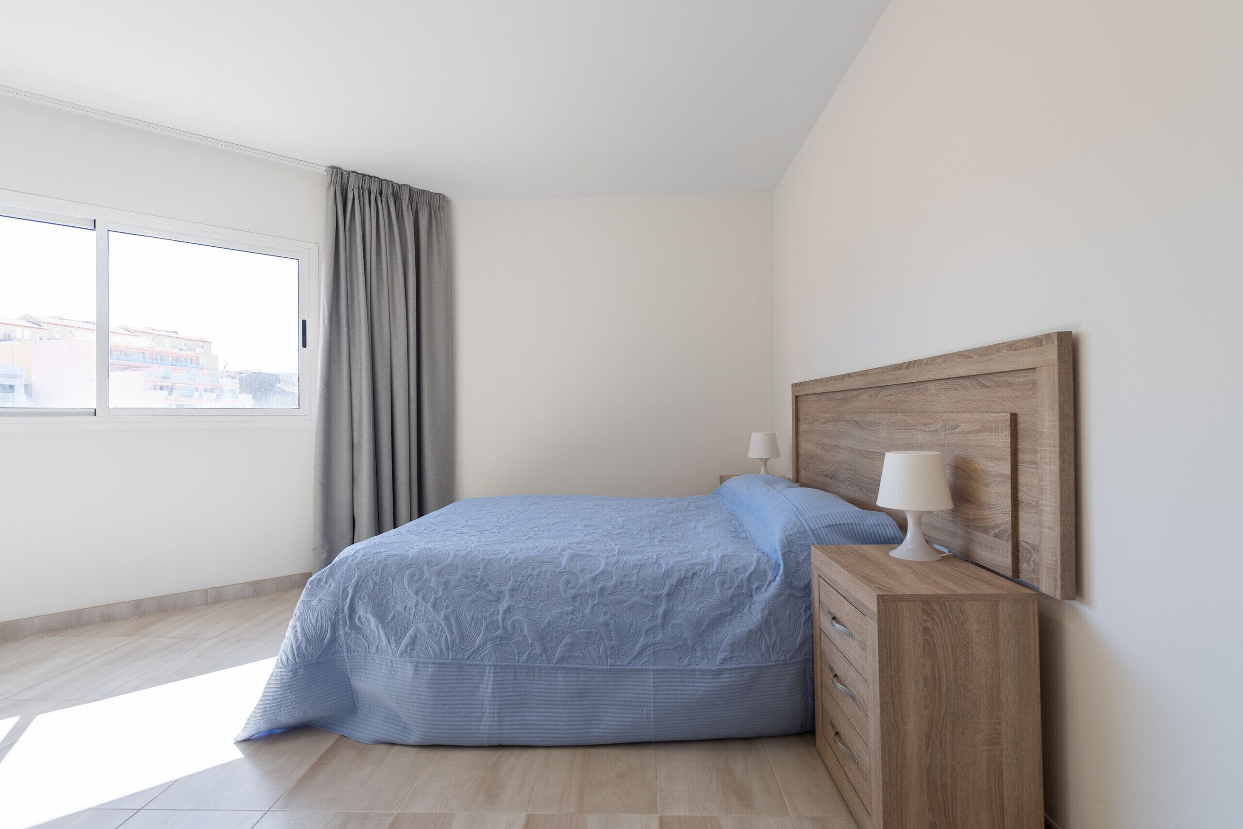 Ein Schlafzimmer mit einem Bett, einem Nachttisch, einer Lampe und einem Fenster mit Vorhängen.