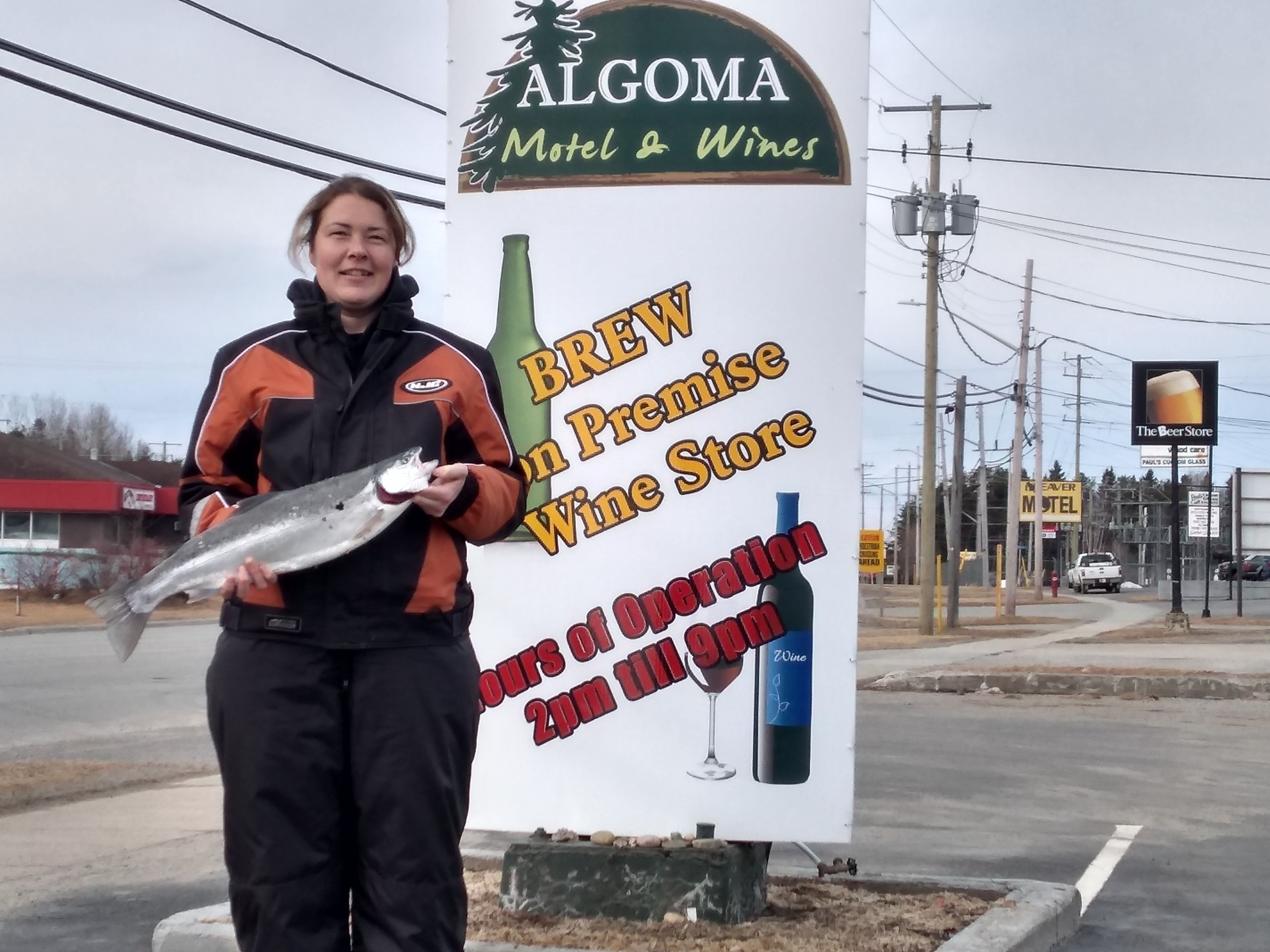 A person holding a fish in front of a sign for Algoma Motel & Wines, which advertises Brew on Premise Wine Store and hours of operation until 2pm.
