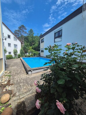 En swimmingpool omgivet af en belagt terrasse med en hvid bygning og en have med lyserøde blomster.