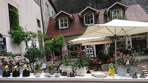 En hyggelig udendørs café med mange krukker med planter og blomster, en lille butik i baggrunden og en rustik bygning med skråt tag.