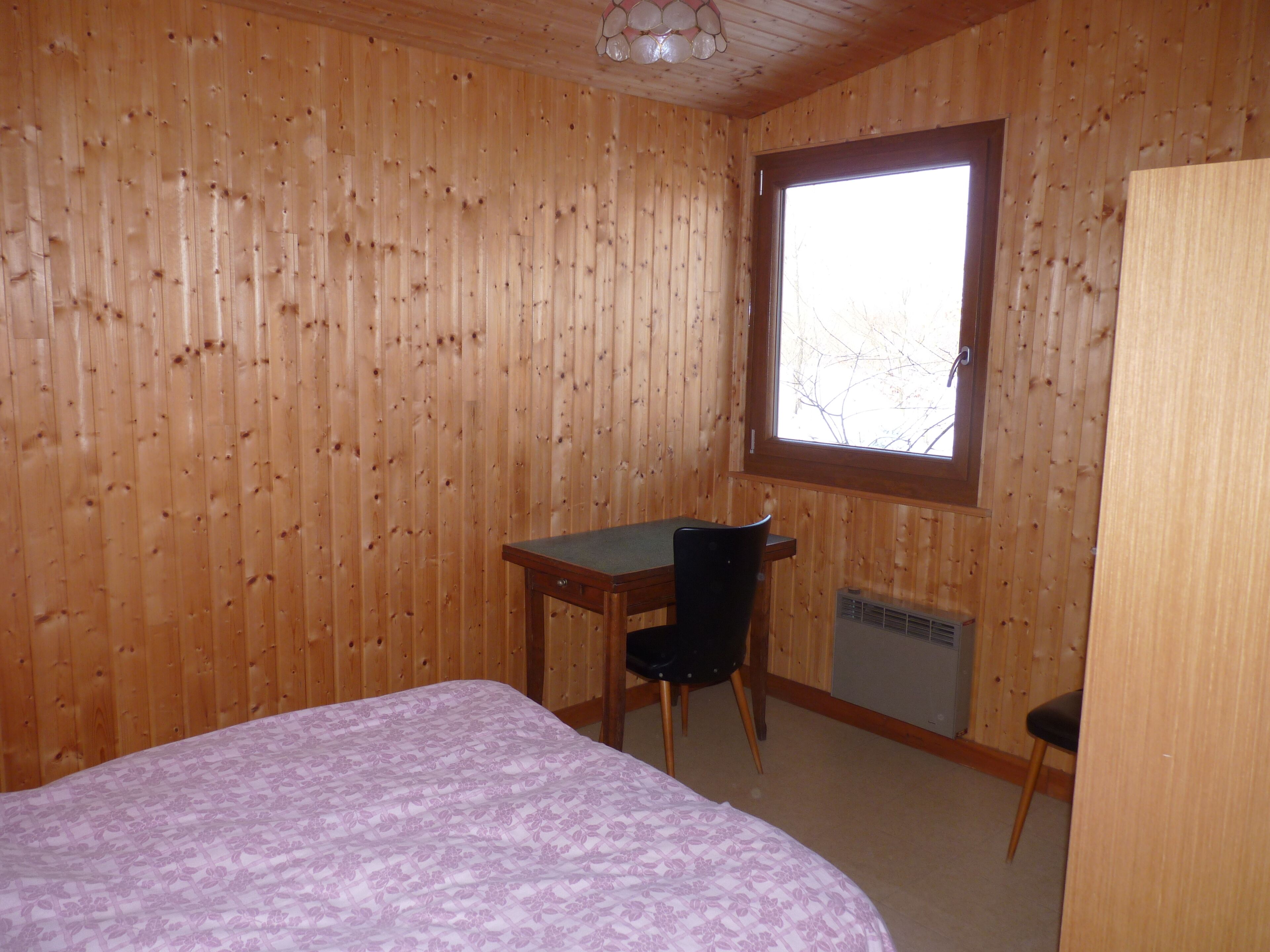 A room with wooden walls, a bed with a floral bedspread, a desk with a chair, and a window with a view of bare trees.