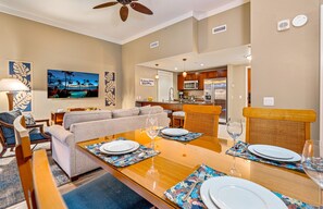 A modern living room with a sofa, dining table set for two, and a kitchen in the background. - Maui Resort Rentals: Honua Kai Hokulani 140 – Upgraded Large Ground Floor 1BR, Spacious Lawn Area + Easy Pools/Beach Access (Lahaina)