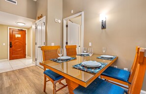 A dining area with a glass-top table, blue cushioned chairs, and a wall-mounted light fixture. - Maui Resort Rentals: Honua Kai Hokulani 140 – Upgraded Large Ground Floor 1BR, Spacious Lawn Area + Easy Pools/Beach Access (Lahaina)