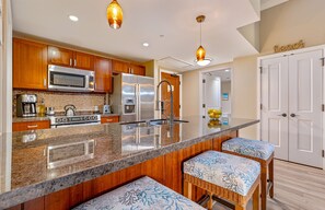A kitchen with wooden cabinets, stainless steel appliances, a granite countertop, and two wicker bar stools. - Maui Resort Rentals: Honua Kai Hokulani 140 – Upgraded Large Ground Floor 1BR, Spacious Lawn Area + Easy Pools/Beach Access (Lahaina)