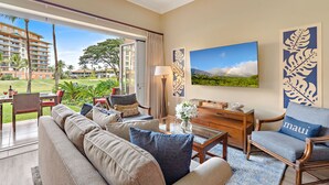 Una camera d'albergo con una grande finestra, un divano, un tavolo da pranzo e una sedia. Presente un quadro appeso alla parete e un cuscino decorativo con la scritta "Maui".