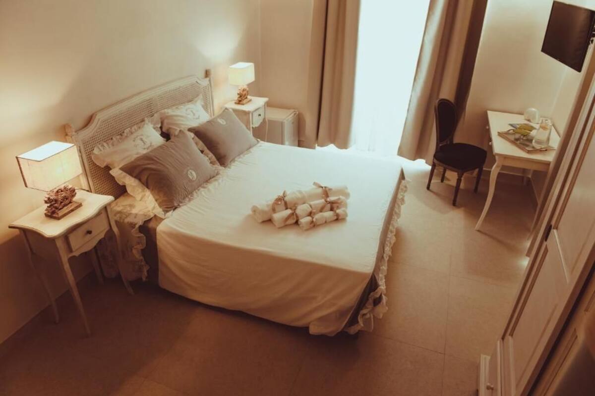 Ein ordentlich bezogenes Bett mit weißer Decke und Kissen, ein Nachttisch mit Lampe, ein Stuhl und ein kleiner Tisch mit Buch und Tasse.