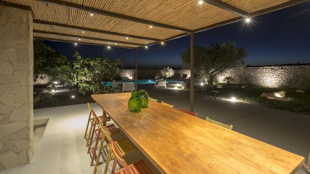 Ein Essbereich im Freien mit Holztisch und Stühlen, Steinwänden und Blick auf einen Pool sowie eine Steinsäule bei Nacht.
