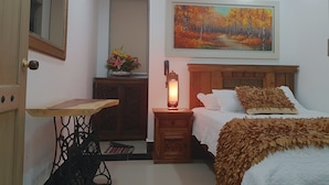 A bedroom with a bed, a nightstand with a lamp, a wooden headboard, a painting on the wall, a mirror, and a small table with a vase of flowers. - Hotel Boutique Campanario del Río (Soledad)
