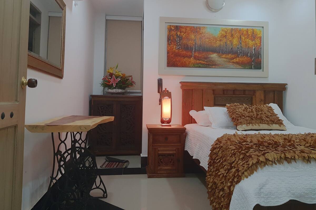 A bedroom with a bed, a nightstand with a lamp, a wooden headboard, a painting on the wall, a mirror, and a small table with a vase of flowers.