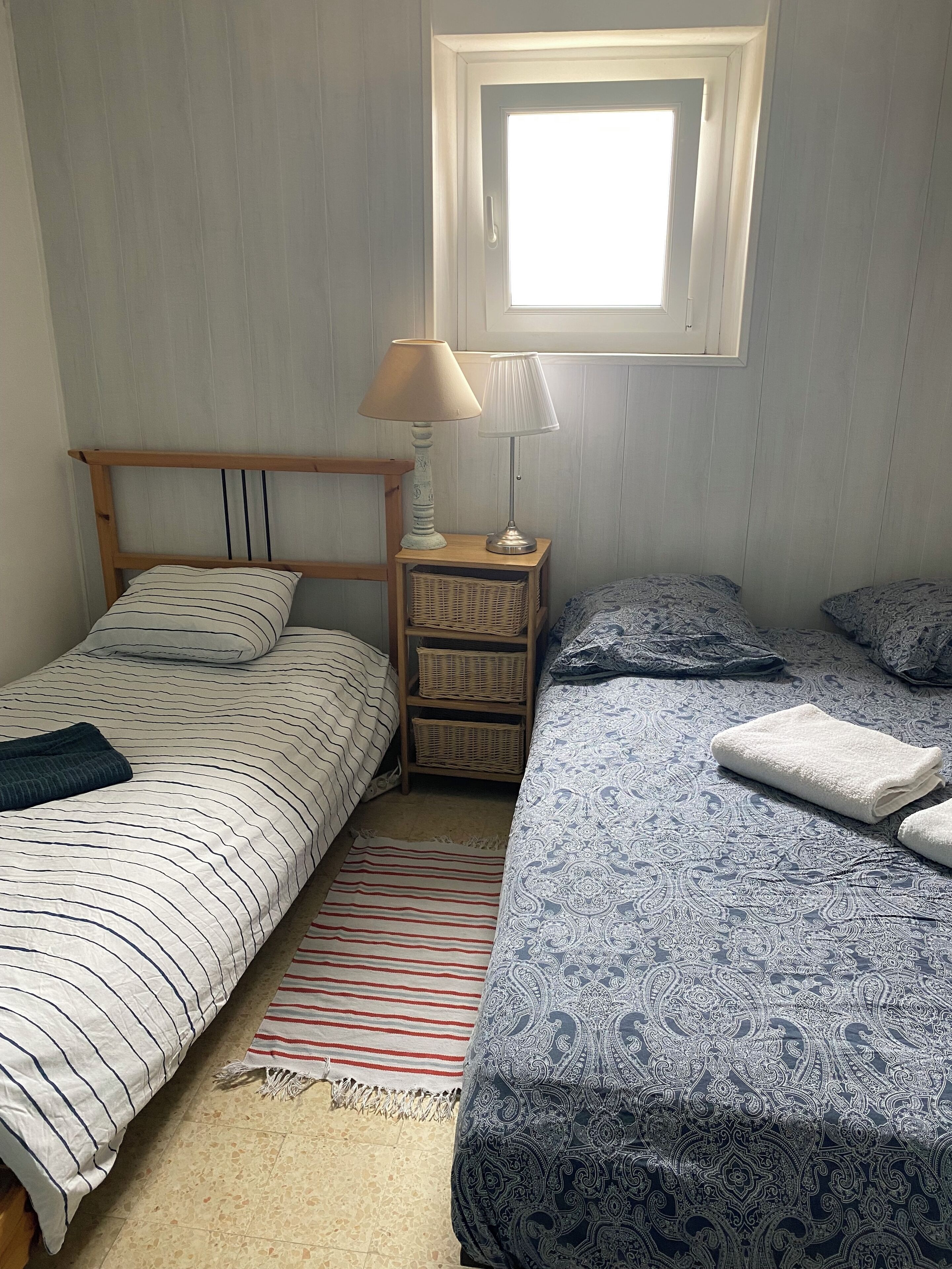 Une chambre avec deux lits, une fenêtre, une table de chevet avec une lampe et un panier en osier.