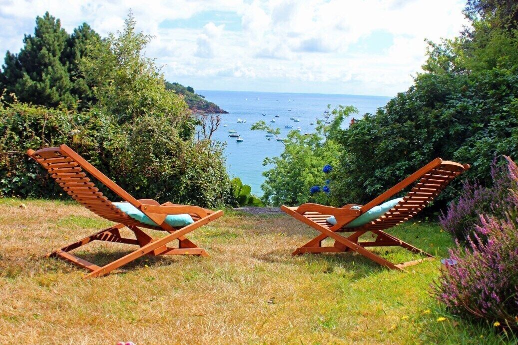 Two wooden lounge chairs facing a coastal view with boats and greenery.