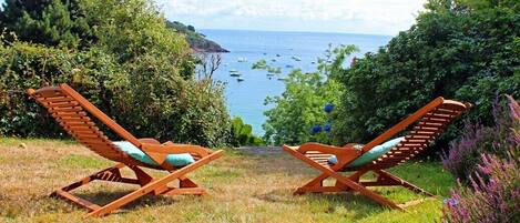 Two wooden lounge chairs facing a coastal view with boats and greenery.