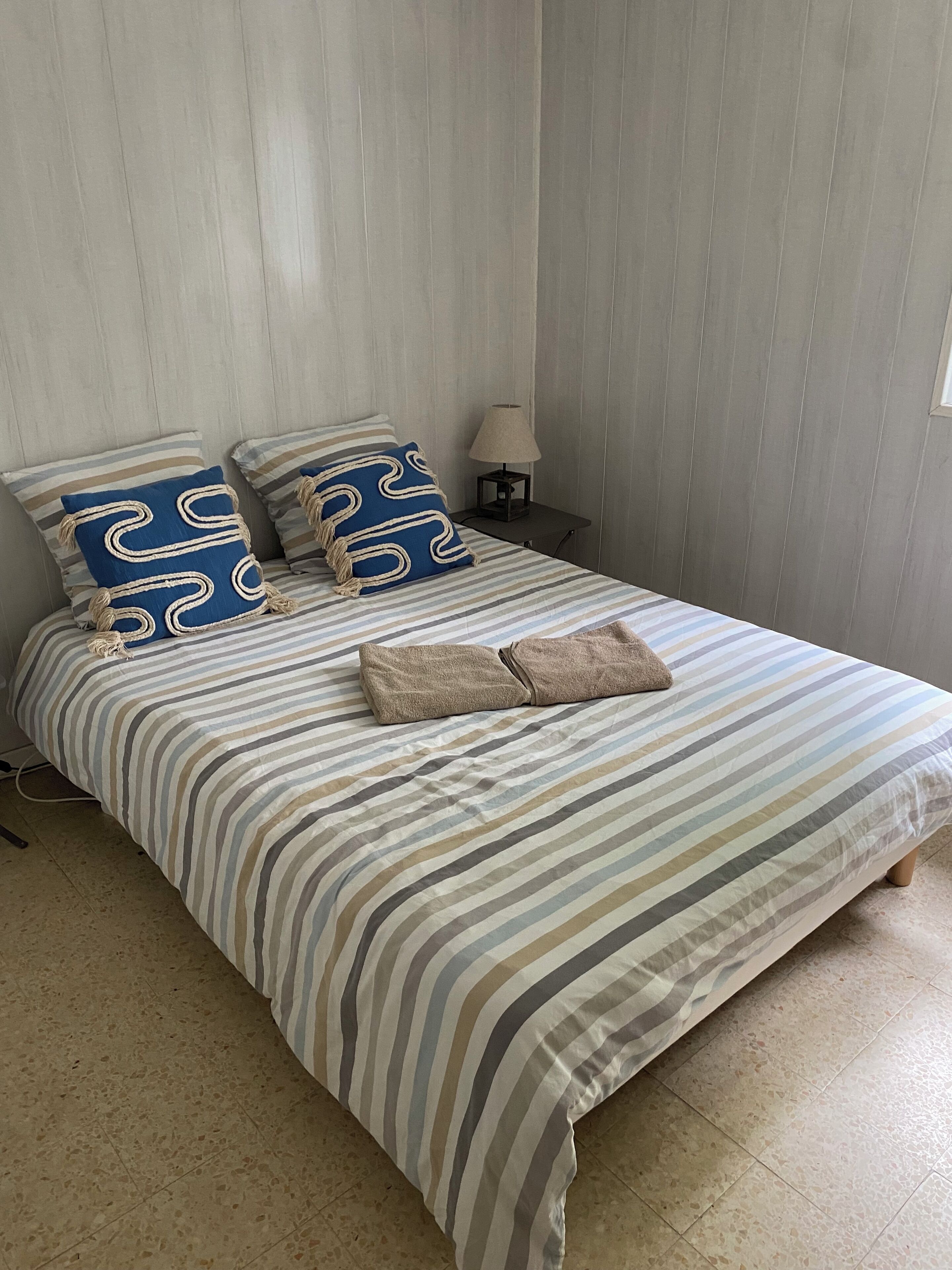 A bed with striped bedding, two pillows with a blue and beige pattern, a small bedside table with a lamp, and a window on the right.