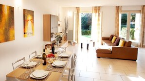 A dining area with a wooden table set for four, a sofa in the living room, and a view of greenery outside. - Tremezzo Residence 2 bedroom home with pool and spa by Italian Apartments
 (Tremezzo)