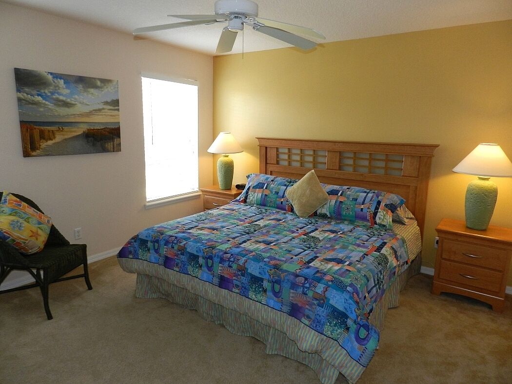 A bedroom with a bed, two nightstands, a chair, a ceiling fan, and a painting on the wall.