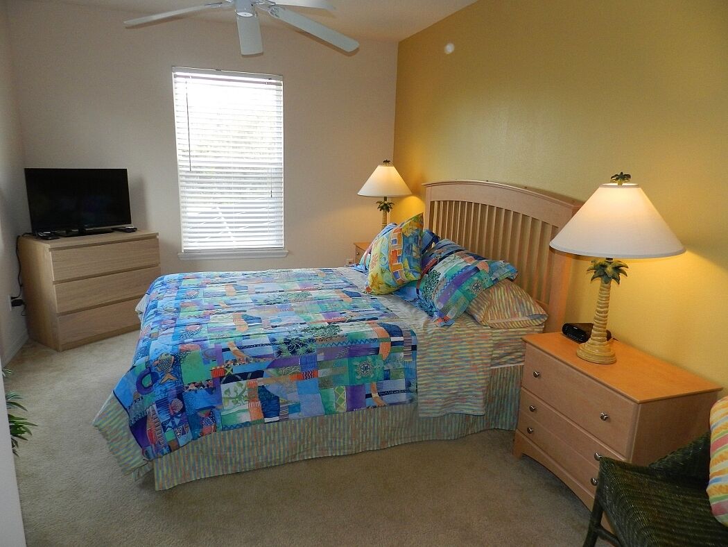 A bedroom with a bed, two bedside lamps, a nightstand, a TV, and a window with blinds.