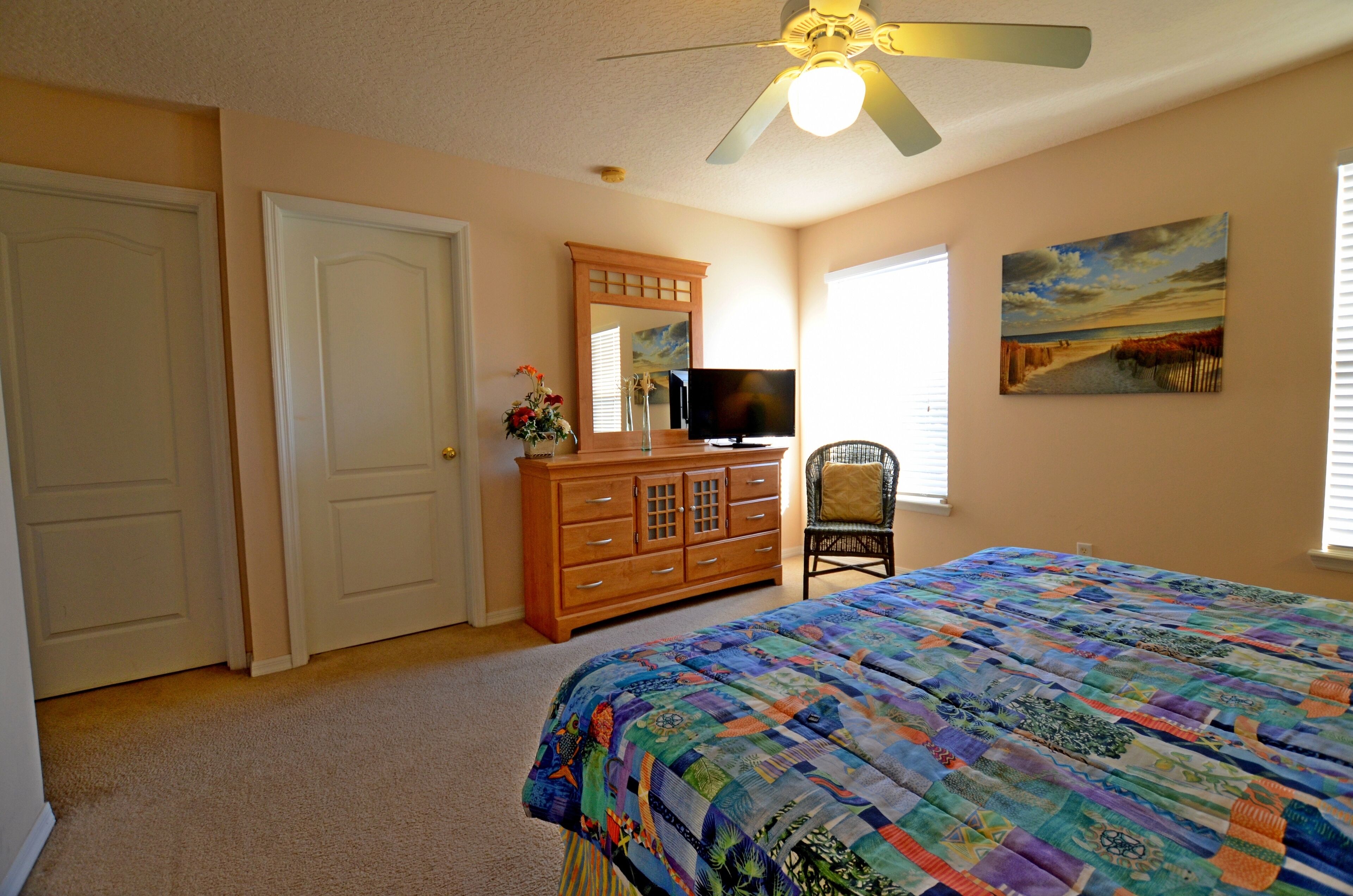 A bedroom with a bed, dresser, chair, TV, and artwork on the wall.