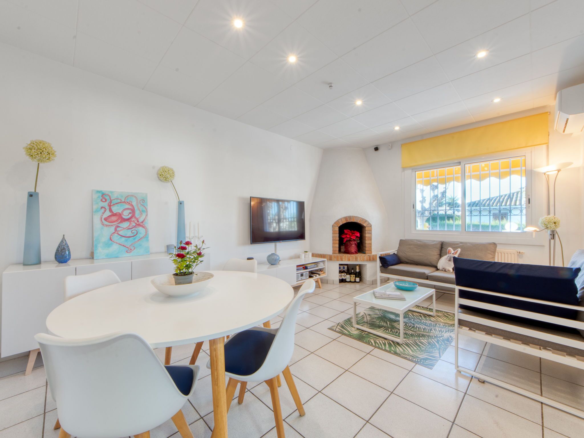 A modern living room with a round dining table, white chairs, a sofa, a TV, and a fireplace.