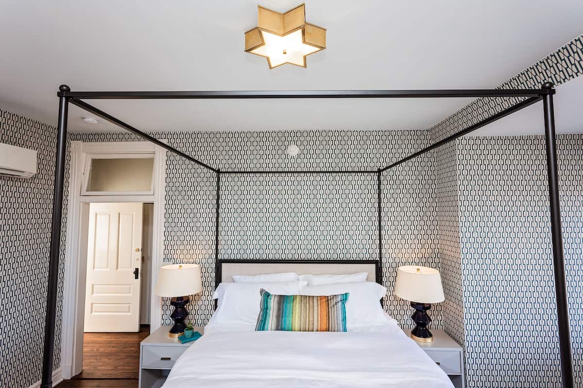 A four-poster bed with white linens and a colorful pillow, two bedside lamps, and a star-shaped ceiling light.