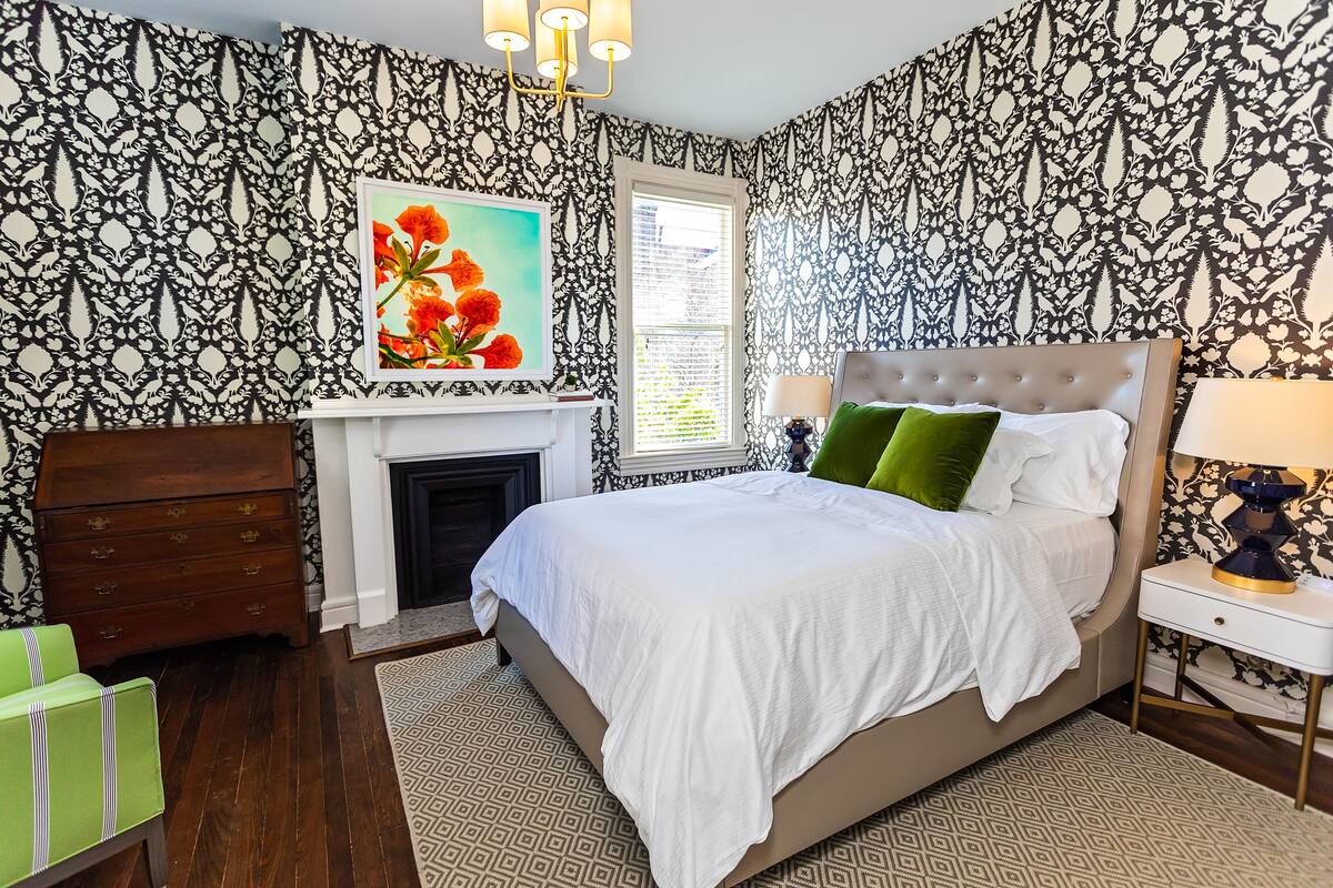 A bedroom with a patterned wallpaper, a bed with white and green bedding, a dresser, a nightstand with a lamp, and a fireplace.