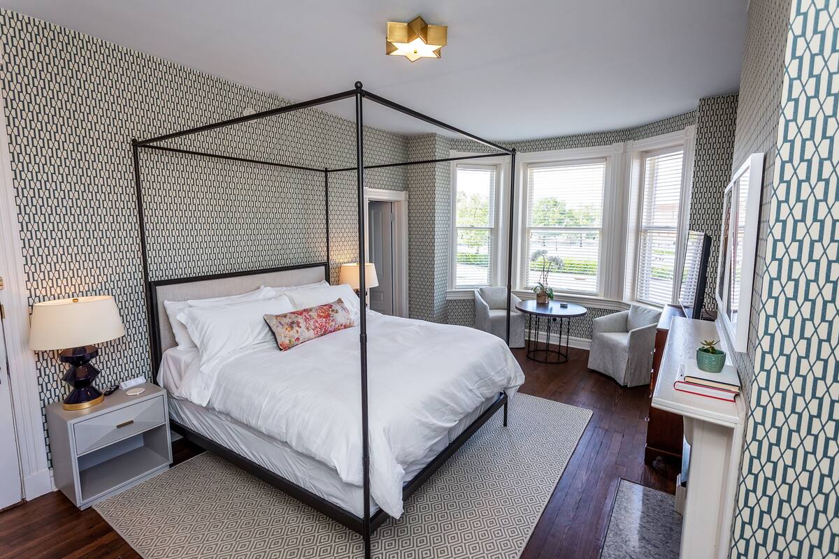 A four-poster bed with a nightstand, a bedside lamp, and a small table with a chair in a bedroom with patterned wallpaper and large windows.