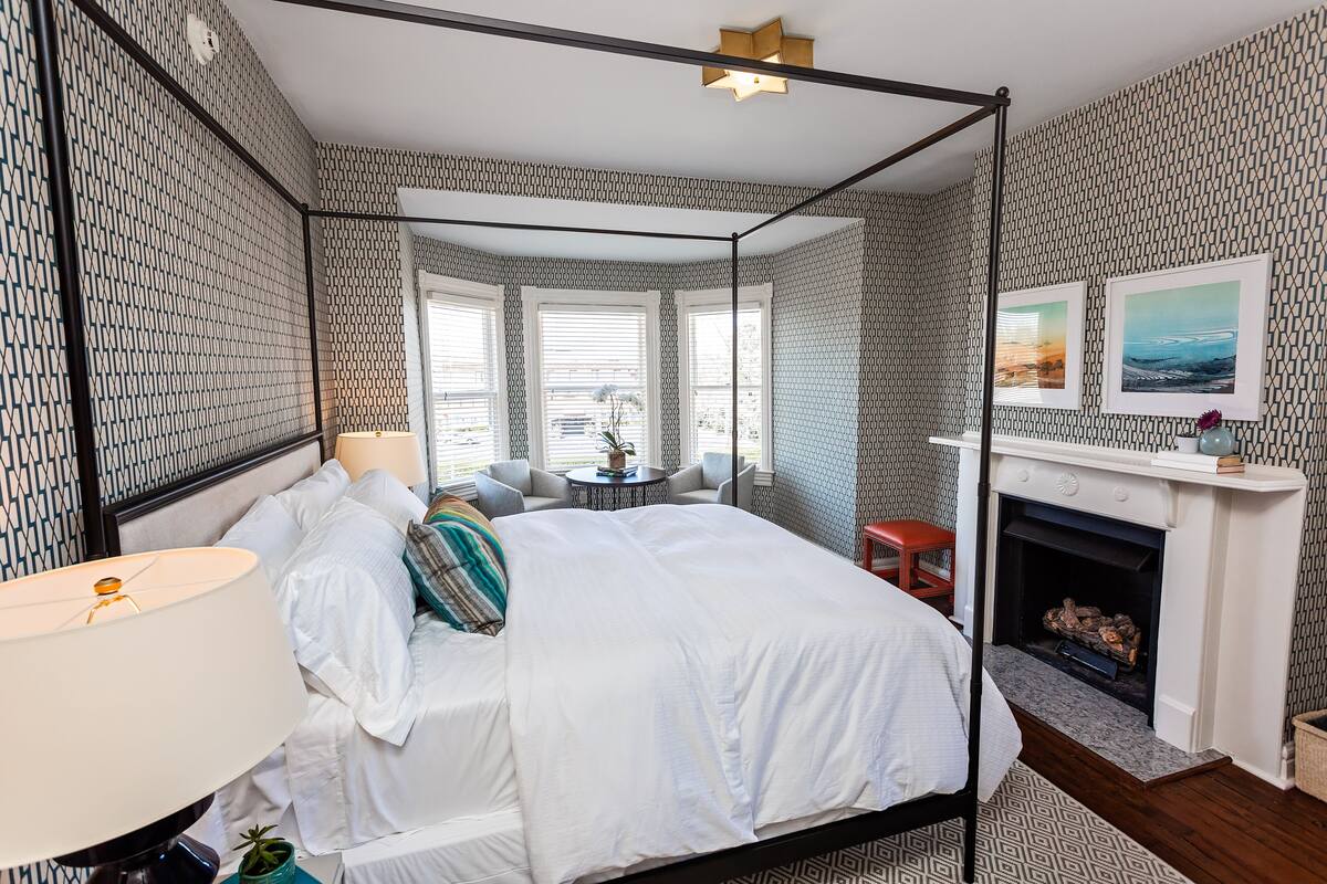 A four-poster bed with white linens, a bedside lamp, and a small round table with a plant. There is a fireplace with a mantel and two framed pictures above it.
