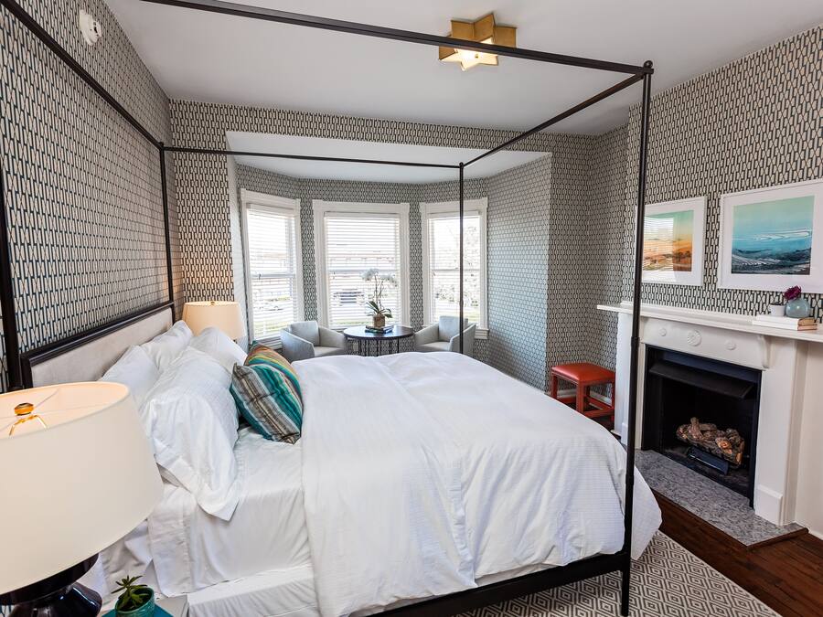 A four-poster bed with white linens, a bedside lamp, and a small round table with a plant. There is a fireplace with a mantel and two framed pictures above it.