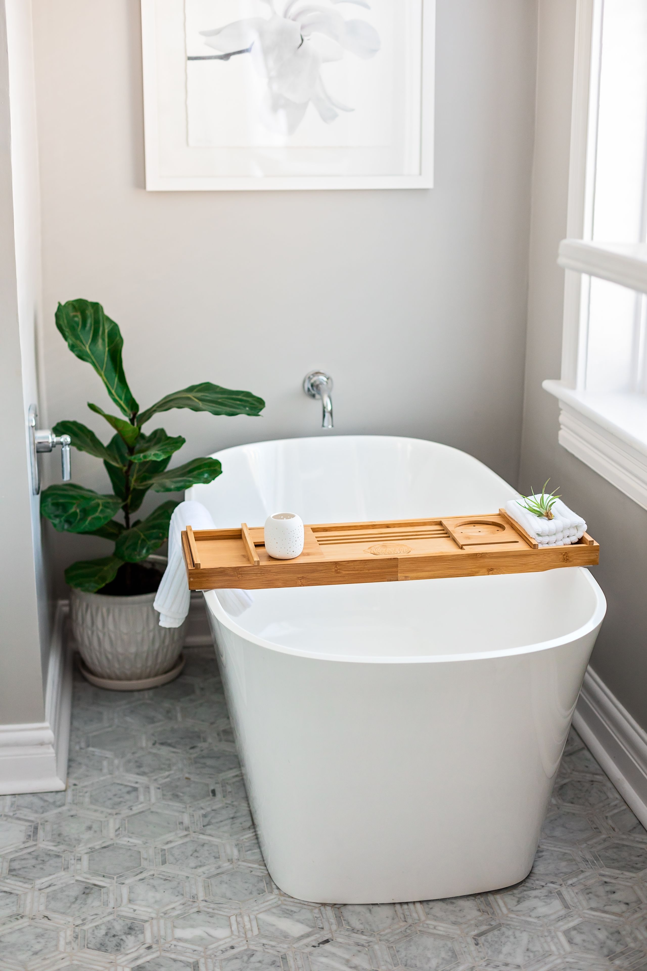 Een moderne badkamer met een vrijstaand bad, een houten dienblad met toiletartikelen, een potplant en een lijst met een kunstwerk aan de muur.