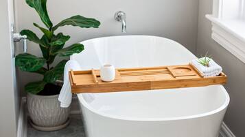 Een moderne badkamer met een vrijstaand bad, een houten dienblad met toiletartikelen, een potplant en een lijst met een kunstwerk aan de muur.