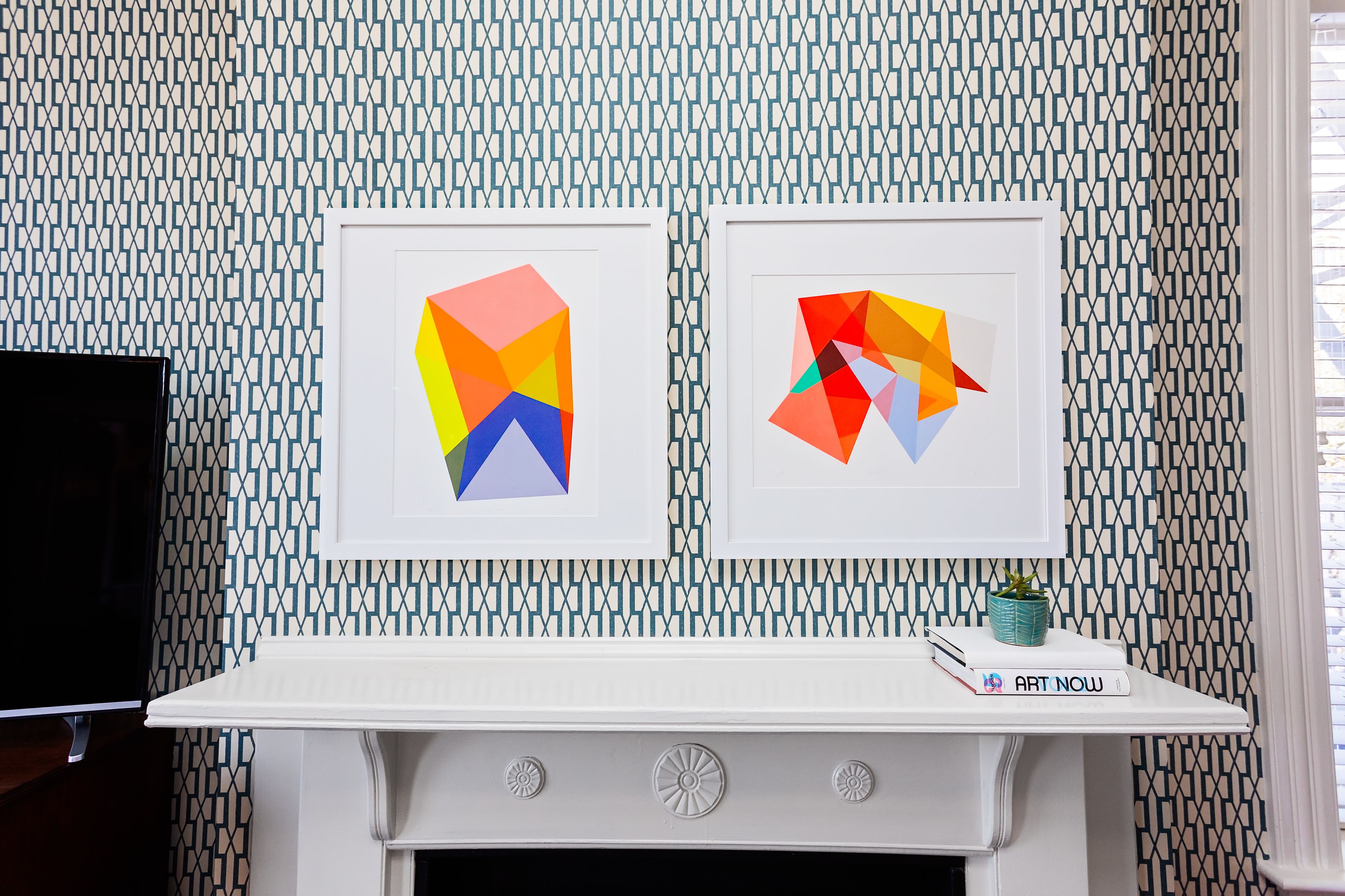 Una habitación con papel tapiz estampado, una chimenea blanca y dos cuadros geométricos enmarcados.
