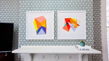 Una habitación con papel tapiz estampado, una chimenea blanca y dos cuadros geométricos enmarcados.