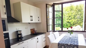 Une cuisine moderne avec des meubles blancs, un plan de travail noir et une machine à café. On y trouve une table à manger recouverte d’une nappe à motifs bleus et blancs, ainsi qu’un vase rempli de fleurs.