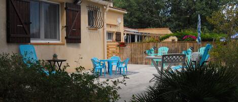 A house with a patio area featuring blue chairs and a table, surrounded by greenery.