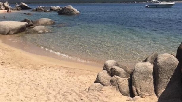 Una playa con agua cristalina, rocas grandes y montañas al fondo.