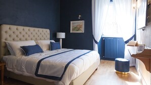 A bedroom with a tufted headboard, a bed with white and blue bedding, a bedside lamp, a framed picture on the wall, a window with curtains, a radiator, and a wooden side table with a lamp. - Villa Klaudia - charming house_ Pompeii (San Giorgio a Cremano)