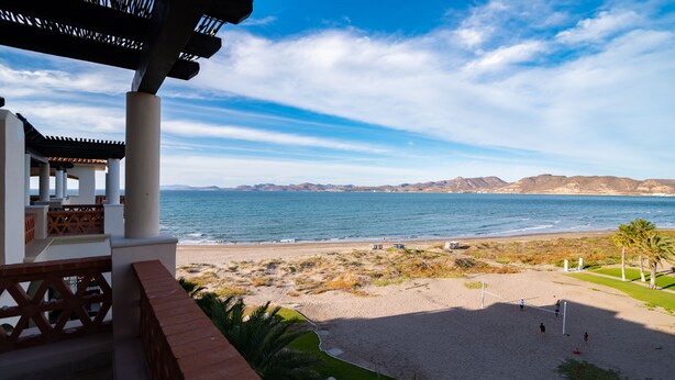 Vista a la playa desde un balcón con pérgola, una playa arenosa y montañas al fondo.