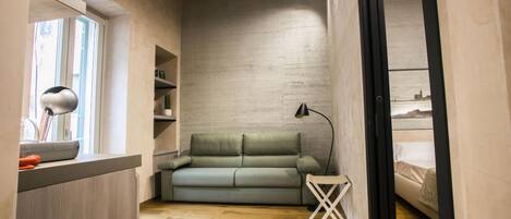 A compact room with a wooden floor, a green sofa, a small white chair, and a desk with a lamp.