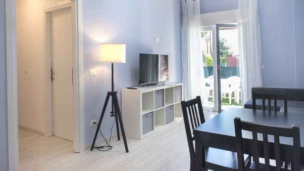 A room with a dining table, chairs, a TV on a stand, and a lamp. - S051 - Sirolo, new three-room apartment with garden (Sirolo)