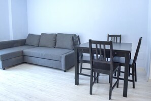 A modern living room with a grey sectional sofa, a wooden dining table with four chairs, and light wooden flooring.