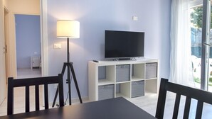 A room with a TV on a white stand, a lamp, and a table with chairs. - S051 - Sirolo, new three-room apartment with garden (Sirolo)