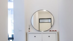 A white cabinet with a round mirror above it, a small shelf with decorative items, and a door with a frosted glass panel. - S051 - Sirolo, new three-room apartment with garden (Sirolo)