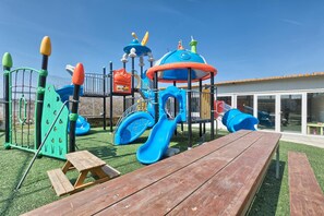 Children can play safely on artificial grass with slides and climbing elements, surrounded by picnic tables for supervision.
