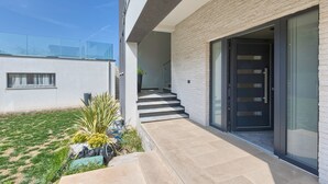 Un bâtiment moderne doté d’un balcon en verre, d’un escalier menant à une porte sombre et d’un petit espace jardin avec des plantes.