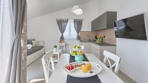 Une cuisine moderne avec des meubles blancs, une table à manger sur laquelle se trouve un plateau de fruits et de jus, et un téléviseur fixé au mur.