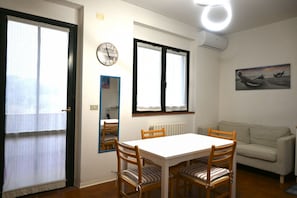 A dining area with a white table and wooden chairs, a clock on the wall, and a painting above a window.