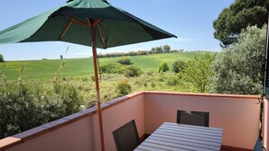 Un balcón con mesa y sillas, un parasol verde y vistas a un campo y árboles.